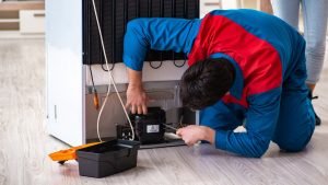 A man kneels on the floor, focused on repairing a refrigerator, surrounded by tools and parts.
